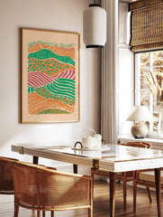 Illustrative abstract landscape print of layered farm fields in orange, green, and purple hues with, in a styled room mockup