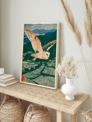 Barn owl in flight over terraced fields and mountains, green, blue, beige woodblock print, in an alternate framed mockup