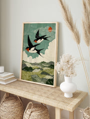 Two barn swallows flying over green hills under a teal sky with white clouds and red sun in, in an alternate framed mockup