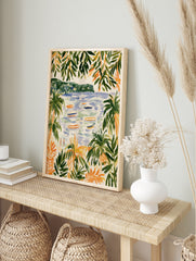 Coastal beach scene with palm trees, boats on water, and distant hills, illustrative art print, in an alternate framed mockup
