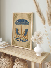 Illustrative beach scene art print with blue and white striped parasol and two deck chairs on, in an alternate framed mockup