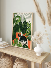 Black and white cat sitting among large green house plants in terracotta pots, botanical, in an alternate framed mockup