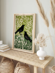 Linocut style print of two black birds amidst green and white flowers and foliage, in an alternate framed mockup