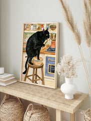 Watercolour illustration of a black cat standing on a kitchen stool tasting from a bowl with, in an alternate framed mockup
