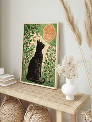 Linocut style art print of a black cat sitting in green foliage, looking at a large orange, in an alternate framed mockup