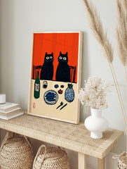 Illustrative print of two black cats at a dinner table with fish, sake, and a vase, against, in an alternate framed mockup
