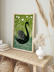 Black swan swimming among white wildflowers and green reeds in a linocut style art print, in an alternate framed mockup