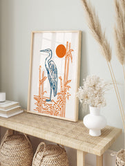 Illustrative blue heron standing by orange bamboo stalks with a vibrant orange sun on a white, in an alternate framed mockup