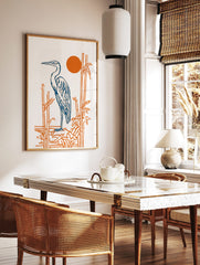 Illustrative blue heron standing by orange bamboo stalks with a vibrant orange sun on a white, in a styled room mockup