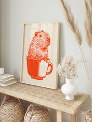 Orange ink illustration of a capybara sitting inside a teacup on off-white paper, in an alternate framed mockup