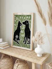 Black cat and dog silhouettes facing each other in green grass with white flowers, linocut, in an alternate framed mockup
