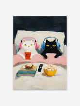 Whimsical cat art print of a white cat and a black cat with headphones enjoying popcorn in bed during a movie night.
