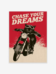 Black cat wearing motorcycle goggles on a bike with red background and the text Chase Your Dreams
