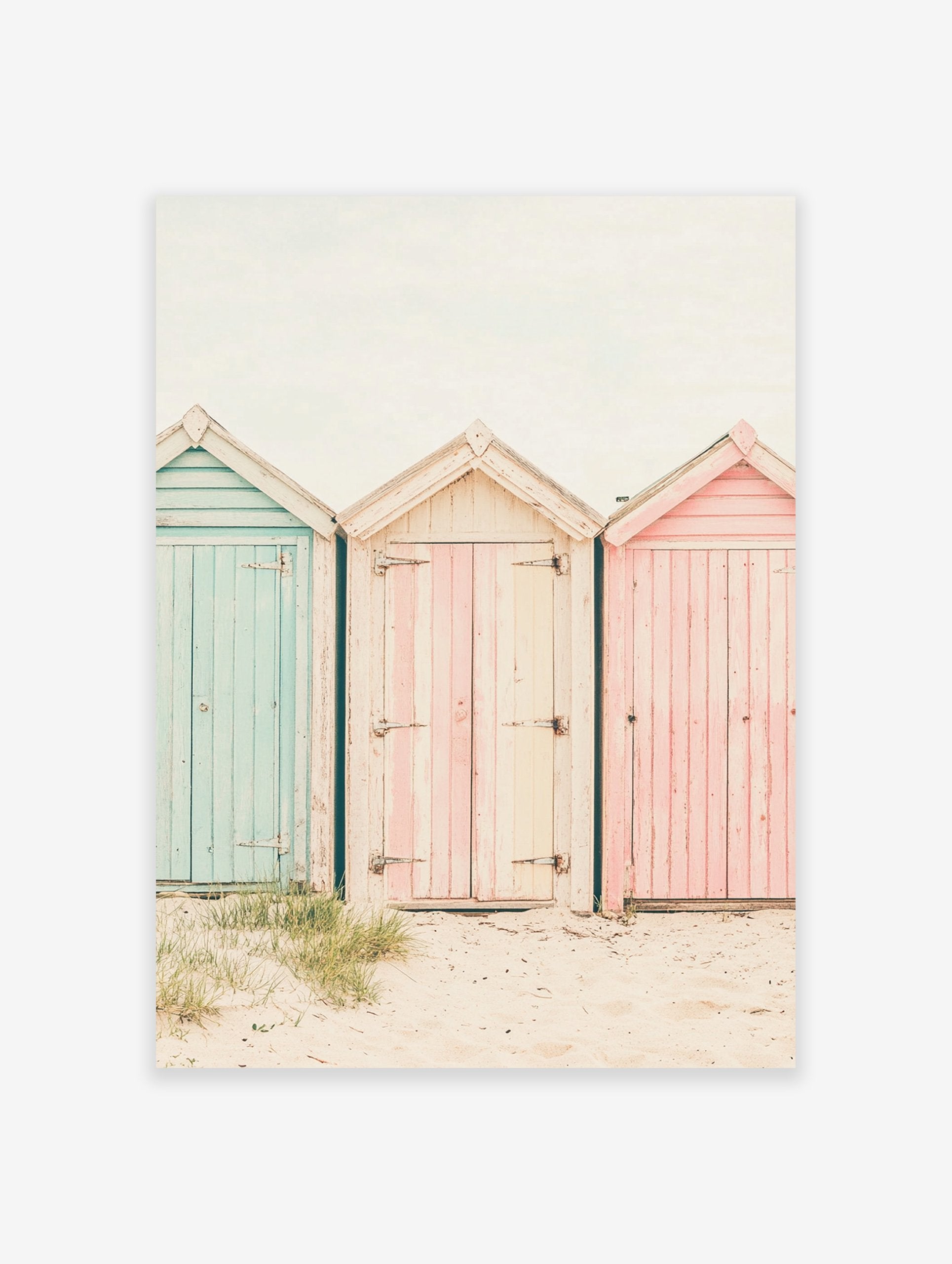 Photography poster of three weathered beach huts in pastel blue, pink, and striped cream on a sandy beach.
