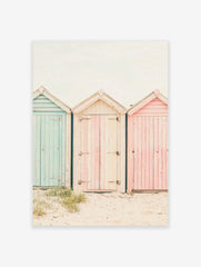 Photography poster of three weathered beach huts in pastel blue, pink, and striped cream on a sandy beach.
