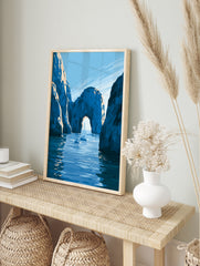 Illustrative coastal wall art print of a natural stone archway over blue water with sailboats, in an alternate framed mockup