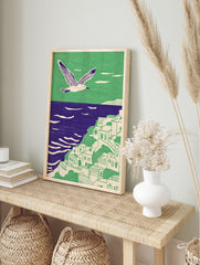 Illustrative coastal wall art with a white seagull flying over a green cliffside village and purple ocean. - Image 3