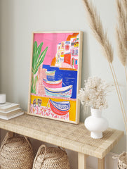 Colourful illustrative coastal town art print with boats in blue water, white buildings, and, in an alternate framed mockup