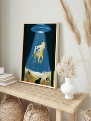 Surreal wall art print of a spotted cow levitating in a bright blue UFO tractor beam within a, in an alternate framed mockup