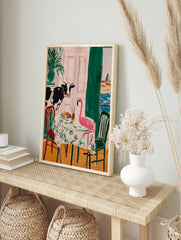 Illustrative wall art of a cow and a pink flamingo dining at a table, with a pink door and, in an alternate framed mockup