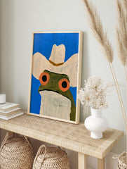 Illustration of a frog wearing a cowboy hat against a bright blue background, art print on, in an alternate framed mockup