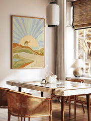 Illustrative desert landscape art print with a camel on a dune, radiating sunset, and distant sea, in a styled room mockup