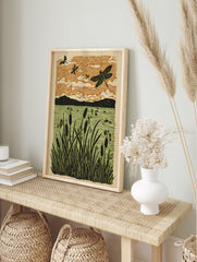 Linocut style art print of dragonflies flying over cattails and water with an orange sky, in an alternate framed mockup