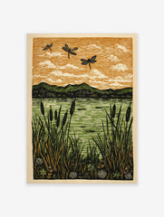 Linocut print of three black dragonflies flying over a green lake with cattails and mountains under an orange sky