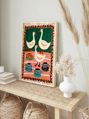 Folk art print of three white ducks, croissants, and coffee cups on green and pink striped, in an alternate framed mockup