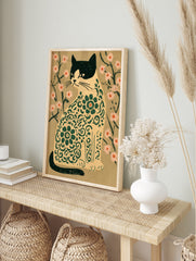 Stylized cat illustration with green floral patterns and pink flowers on a tan background, in an alternate framed mockup