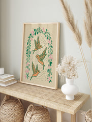 Three orange flying birds within an arched wreath of green leaves and pink flowers on a beige, in an alternate framed mockup