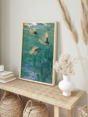 Three flying cranes with red heads and black wing tips over a teal and blue pond with reeds, in an alternate framed mockup