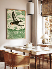 Linocut style print of a duck flying over a marsh with reeds and water, in green, cream, black, and, in a styled room mockup