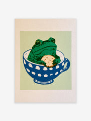 Whimsical green frog sitting in a blue and white polka-dot teacup holding a biscuit on a light green background