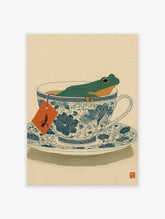 Green frog relaxing in a blue and white patterned teacup, with an orange tea bag tag, on a light beige background.