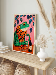 Illustrative wall art of a green frog in a polka-dot scarf, holding a wine glass on a pink, in an alternate framed mockup