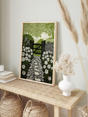 Linocut style print of a garden path leading to a wooden gate, flanked by white roses and, in an alternate framed mockup