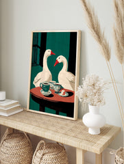 Illustrative wall art of two white geese with red beaks at a table with coffee cups and cake, in an alternate framed mockup