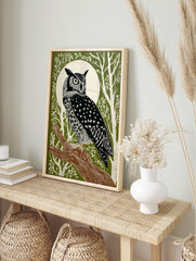 Illustrative great horned owl perched on a branch against a white full moon and green forest, in an alternate framed mockup