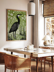 Illustrative linocut print of a black heron with white head markings, standing in green marsh water, in a styled room mockup