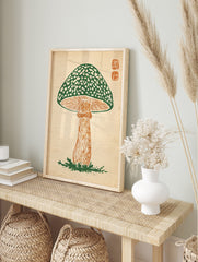 Green mushroom woodcut print with spotted cap, orange stem, and two square seals in the top, in an alternate framed mockup