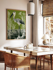 Green linocut print of a patterned stork wading in water with reeds and reflections, in a styled room mockup