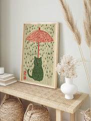 Illustrative green striped cat under a red umbrella with green raindrops on a cream background, in an alternate framed mockup