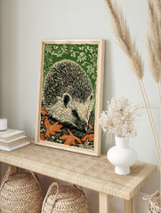 Linocut print of a hedgehog with black quills, off-white face, orange autumn leaves, and, in an alternate framed mockup