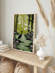 Linocut-style print of a black heron standing in a green forest stream with white flowers and, in an alternate framed mockup
