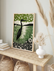 Linocut print of a black heron by a green lake, surrounded by white wildflowers and distant, in an alternate framed mockup