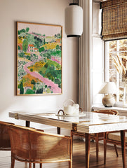 Illustrative landscape art print with a white house, orange roof, green fields, and a winding pink, in a styled room mockup