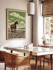 Brown horse standing in tall grass field with green sky and white clouds, linocut style art print, in a styled room mockup