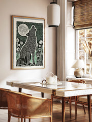 Linocut style art print of a howling wolf with a full moon and flowers in black, white, and green, in a styled room mockup