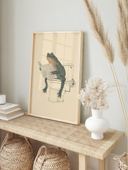 Humorous illustration of a dark green and orange toad sitting on a white toilet, reading a newspaper. - Image 3